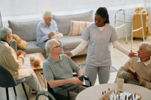 A smiling older adult sits in a cozy lounge, chatting with a caregiver while other residents read nearby.