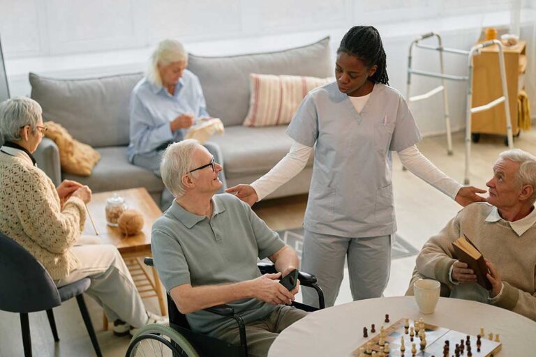 A smiling older adult sits in a cozy lounge, chatting with a caregiver while other residents read nearby.